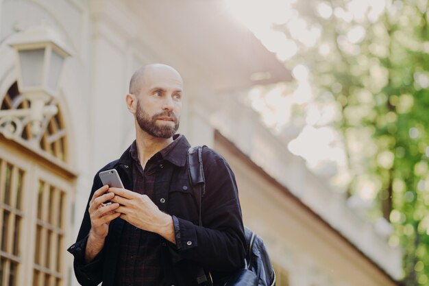 Vue extérieure d'un touriste masculin réfléchi portant un sac à dos tient un téléphone intelligent moderne dans les mains alors qu'il essaie de trouver le bon chemin regarde des cartes en ligne concentrées quelque part