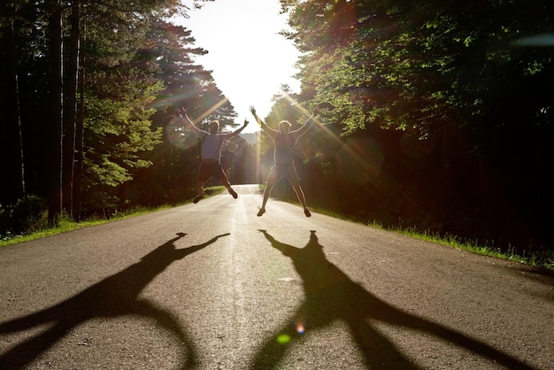 Vue de face d'amis sautant au milieu de la route. Vue horizontale d'hommes sautant dans les ombres du rétroéclairage voyageant dans la nature. Concept de vacances d'été.