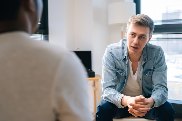 Vue de face d'un beau leader masculin organisant un séminaire de consolidation d'équipe assis en cercle sur une chaise près de la fenêtre. Équipe commerciale multiethnique ayant une réunion de remue-méninges pour planifier et atteindre l'objectif.