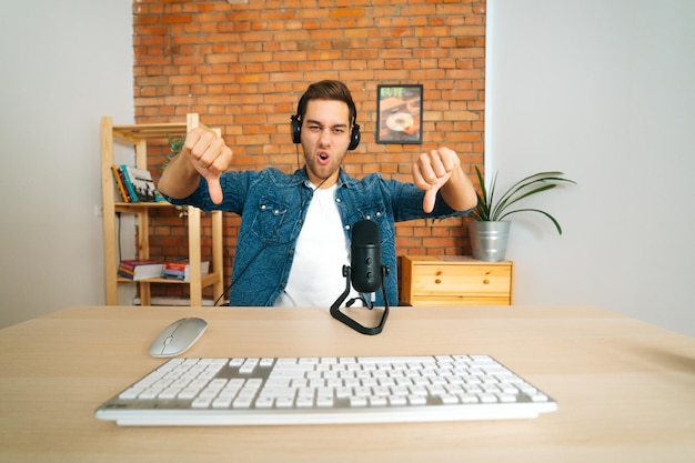 Vue de face d'un blogueur masculin en colère négatif diffusant un podcast en direct à l'aide d'un microphone professionnel montrant deux pouces vers le bas, diffusant au bureau à domicile, regardant la caméra.