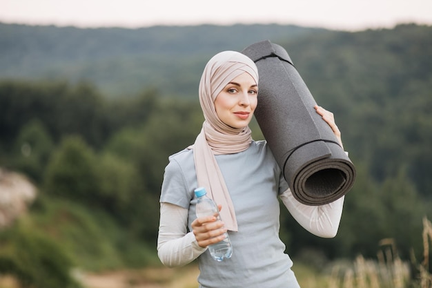 Vue de face d'une charmante femme en hijab et vêtements de sport debout dans un parc verdoyant et tenant un tapis de yoga