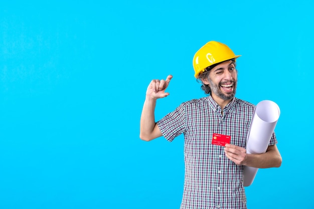 Vue de face constructeur masculin tenant un plan et une carte bancaire sur fond bleu bâtiment d'architecture de travail de constructeur de couleur