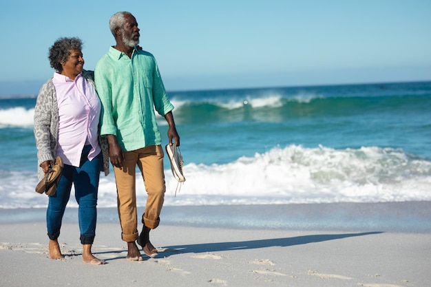 Vue de face d'un couple afro-américain senior marchant sur la plage avec le ciel bleu et la mer en arrière-plan, se tenant la main, marchant pieds nus, portant leurs chaussures et regardant ailleurs