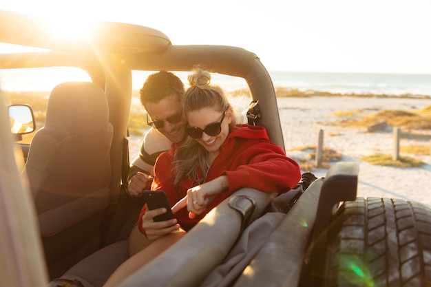 Vue de face d'un couple caucasien à l'intérieur d'une voiture à toit ouvert, utilisant son smartphone et souriant. Week-end vacances à la plage, style de vie et loisirs.