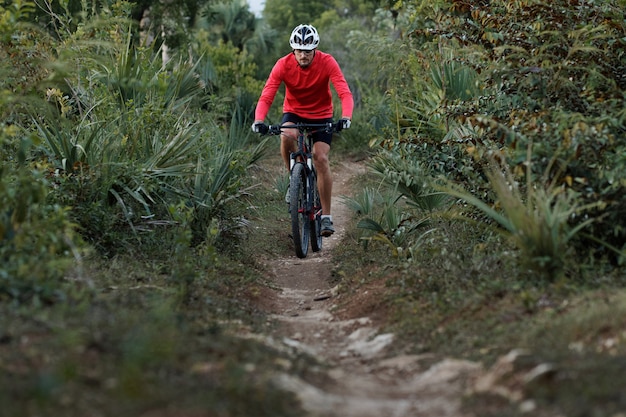 Vue de face d'un cycliste sur un sentier étroit, portant un casque de vélo et un maillot de cyclisme rouge.