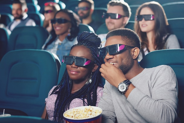 Vue de face de deux amis dans des lunettes 3D assis dans une salle de cinéma moderne, manger du pop-corn et regarder un film drôle.