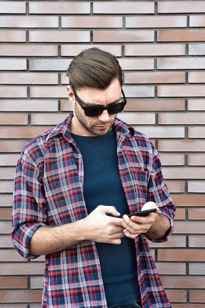 Vue de face du beau jeune homme en tenue décontractée intelligente tenir un téléphone mobile en se tenant debout sur le fond de mur de brique.