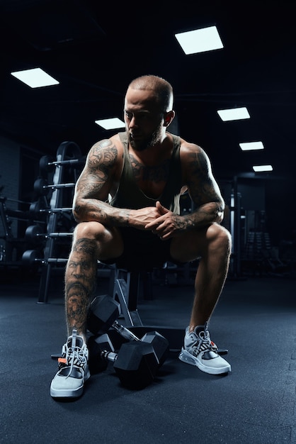 Vue de face du bodybuilder assis sur un banc avec des haltères sur le sol.