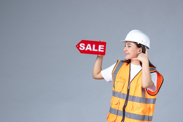 Vue de face du constructeur féminin tenant un tableau de vente rouge sur un mur gris