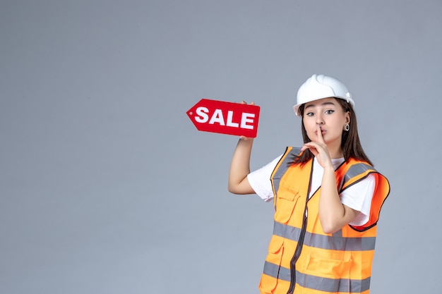 Vue de face du constructeur féminin tenant un tableau de vente rouge sur un mur gris