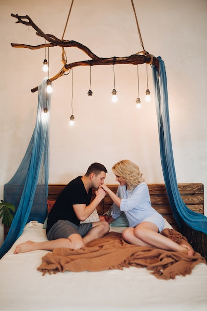 Vue de face du couple assis dans une chambre blanche, étalant du temps romantique ensemble. Beau mari embrasser sa femme blonde dans la main