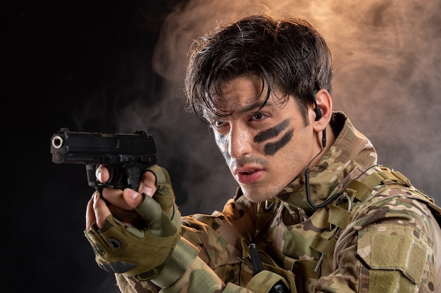 Vue de face du jeune soldat en tenue de camouflage avec pistolet sur mur noir