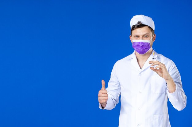 Vue de face du médecin de sexe masculin en costume médical et masque tenant le vaccin et l'injection sur bleu