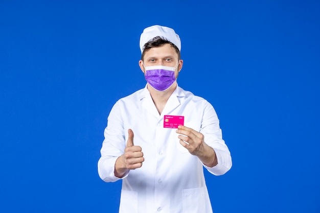 Vue de face du médecin de sexe masculin en costume médical et masque violet tenant la carte de crédit sur bleu