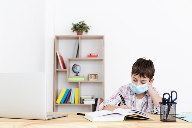 Vue de face de l'enfant faisant ses devoirs tout en portant un masque médical