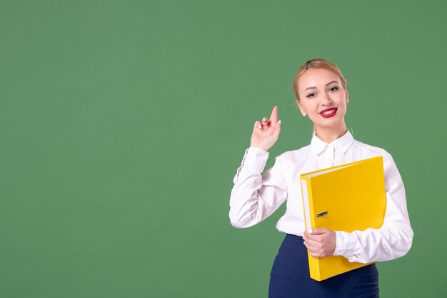 Vue de face enseignante posant avec des fichiers jaunes sur fond vert bibliothèque scolaire étude universitaire uniforme femme livre travail étudiant