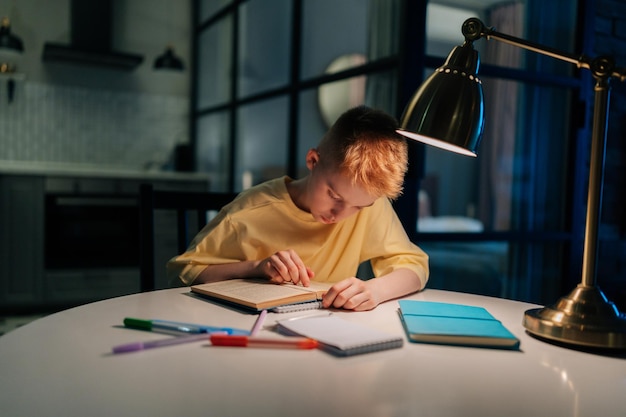 Vue de face d'un étudiant élève concentré qui étudie à la maison en lisant un livre papier faisant ses devoirs en apprenant assis à table sous la lumière d'une lampe la nuit