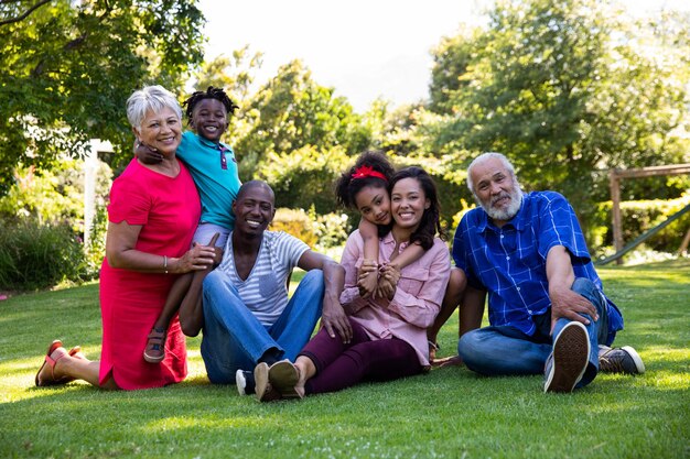 Vue de face d'une famille multiethnique et multigénérationnelle à l'extérieur dans le jardin, assis sur l'herbe ensemble embrassant, regardant la caméra et souriant