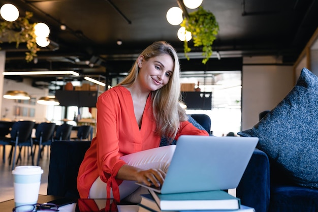 Vue de face d'une femme d'affaires caucasienne aux longs cheveux blonds, portant des vêtements élégants, travaillant dans un bureau moderne, utilisant son ordinateur portable.