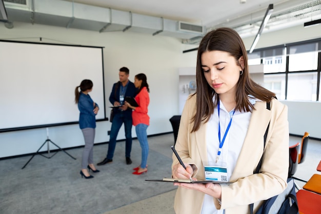 Vue de face d'une femme d'affaires caucasienne travaillant dans un bureau moderne, écrivant sur un presse-papiers, avec des collègues debout à côté d'un tableau blanc discutant ensemble en arrière-plan