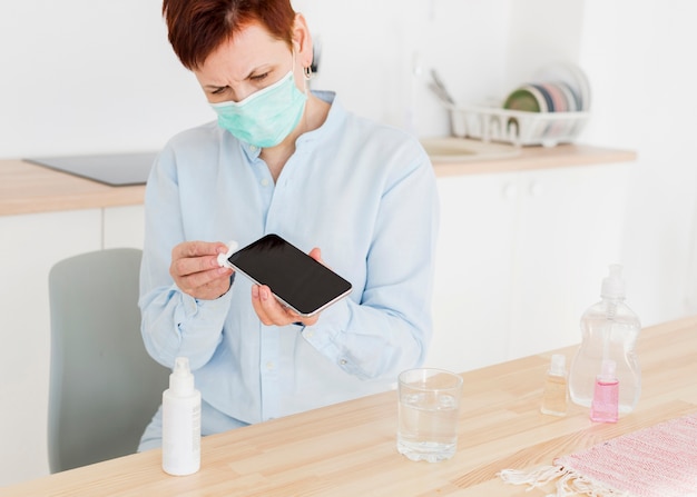 Photo vue de face d'une femme aînée avec un masque médical désinfectant son smartphone à la maison