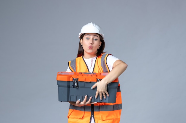 Vue de face d'une femme builder tenant une boîte à outils sur un mur gris