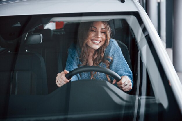 Vue de face d'une femme positive en chemise bleue qui se trouve à l'intérieur d'une nouvelle voiture neuve Dans un salon de l'auto ou à l'aéroport