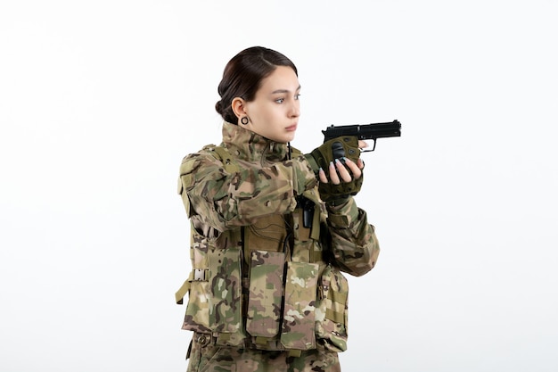 Vue de face femme soldat en tenue de camouflage avec pistolet sur mur blanc