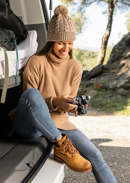 Photo vue de face d'une femme souriante assise dans le coffre de la voiture lors d'un voyage sur la route et tenant la caméra