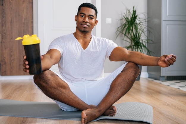 Vue de face d'un homme afro-américain musclé confiant assis les jambes croisées se relaxant sur un tapis d'exercice après l'entraînement et tenant une bouteille d'eau minérale fraîche dans la chambre domestique.