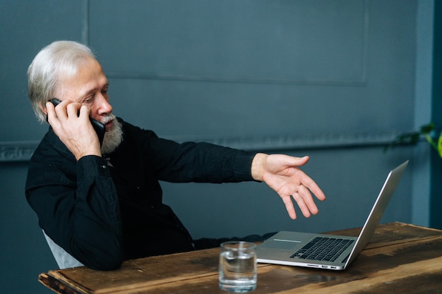 Vue de face d'un homme âgé aux cheveux gris en colère parlant au support technique et demandant de l'aide pour réparer un ordinateur portable cassé assis à une table en bois, au bureau à domicile.