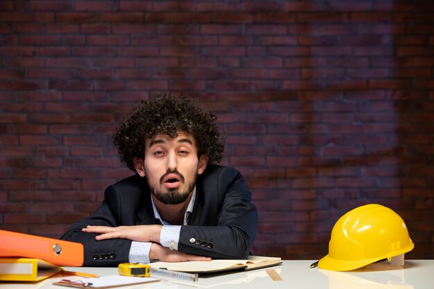 Photo vue de face homme fatigué ingénieur assis derrière le lieu de travail en costume entrepreneur builder plan d'occupation du travail d'entreprise projet d'emploi