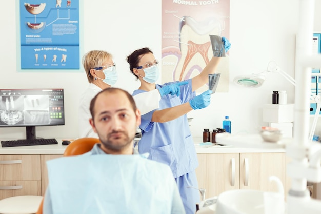 Vue De Face D'un Homme Malade Regardant La Caméra En Attendant Que L'équipe Médicale Examine La Radiographie Somatologique Assise Sur Une Chaise Dentaire Dans Le Bureau De L'hôpital Pour Débiteurs. Stomatologue Prenant Rendez-vous Dentaire