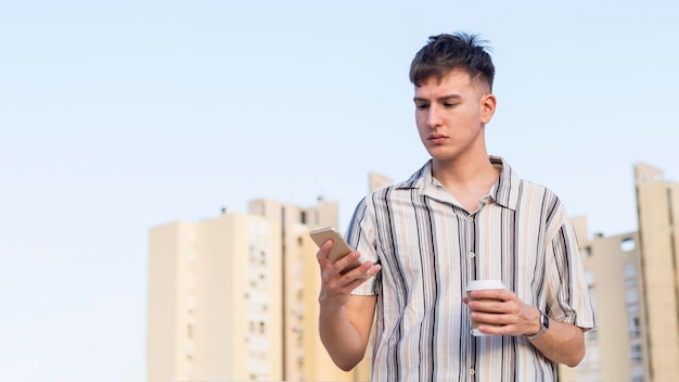 Vue de face de l'homme regardant le smartphone à l'extérieur tout en tenant une tasse de café