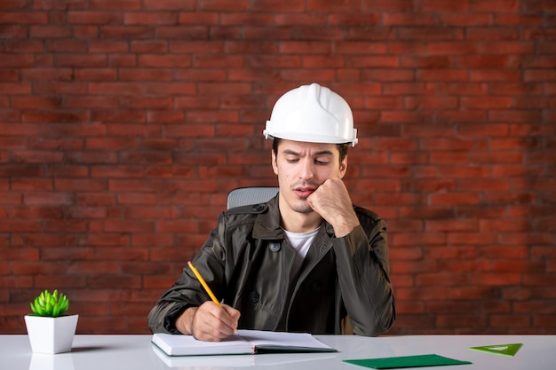 Vue de face ingénieur masculin assis derrière son lieu de travail dans un document de casque blanc propriété d'entreprise plan de constructeur d'agenda de travail