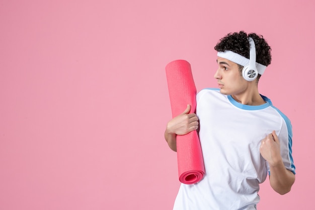 Vue de face jeune athlète masculin en vêtements de sport avec tapis de yoga sur mur rose