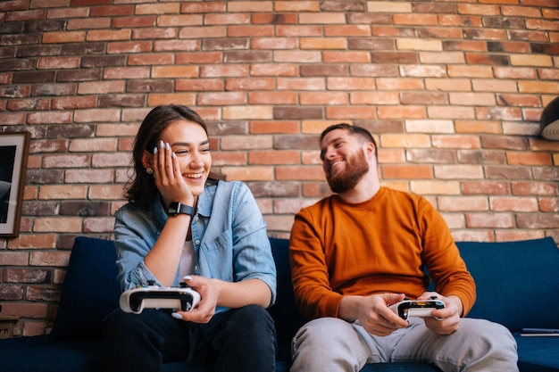 Vue de face d'un jeune couple riant heureux tenant des contrôleurs et jouant à des jeux vidéo sur console assis ensemble sur un canapé dans un salon confortable. Concept d'activité de loisirs des amoureux à la maison.