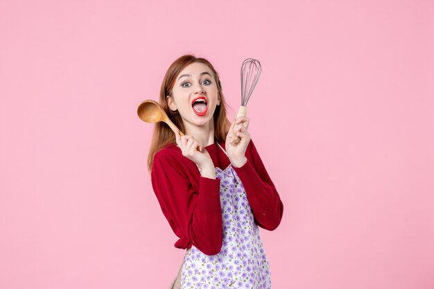 vue de face jeune femme au foyer tenant un fouet et une cuillère en bois sur fond rose pâte à gâteau cuisine cuisson uniforme horizontale profession tarte