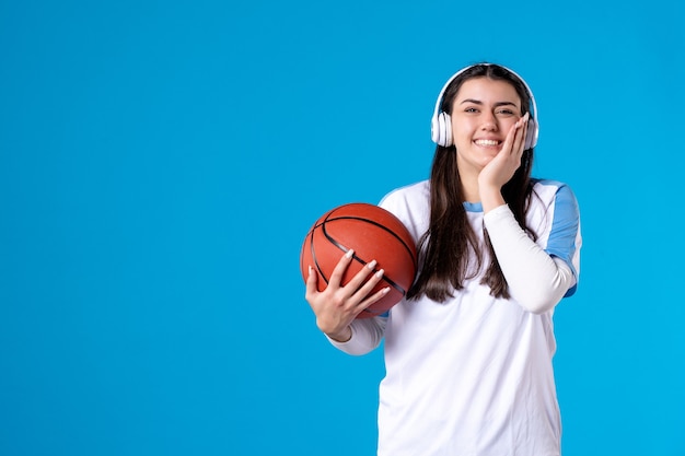 Vue de face jeune femme avec des écouteurs tenant le basket-ball sur le mur bleu