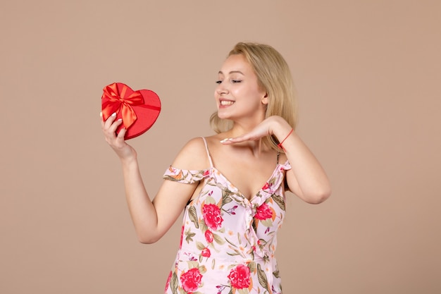 Vue de face d'une jeune femme posant avec un présent en forme de coeur rouge sur un mur marron