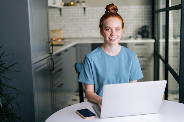 Vue de face d'une jeune femme rousse joyeuse travaillant en tapant sur un ordinateur portable assis à table dans la salle de cuisine. Concept d'activité de loisir femme rousse à la maison pendant l'auto-isolement.