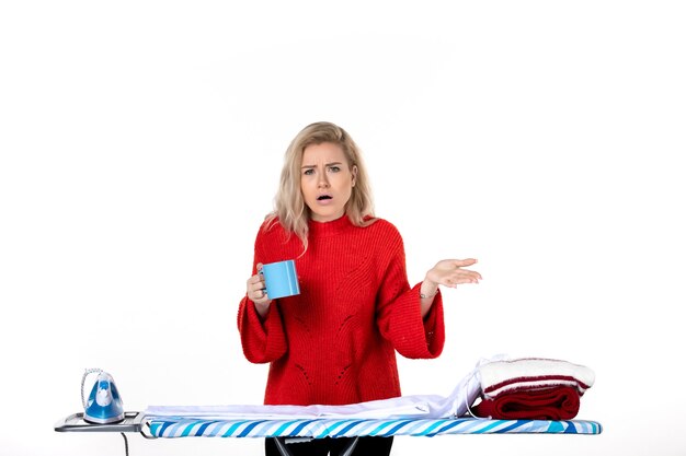 Vue de face d'une jeune femme séduisante curieuse debout derrière la planche à repasser tenant une tasse bleue sur fond blanc