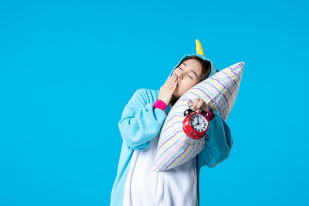 vue de face jeune femme en soirée pyjama tenant des horloges et essayant de s'endormir sur un oreiller sur fond bleu lit rêve sommeil tard repos cauchemar nuit amusement bâillement