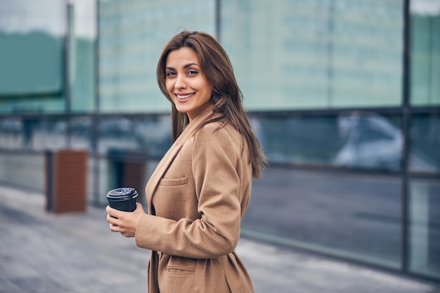 Vue de face d'une jeune femme souriante avec une tasse de café en papier debout à l'extérieur