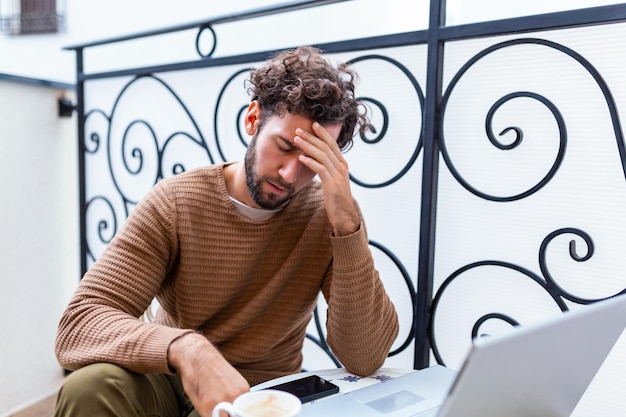 Vue de face. Le jeune homme d'affaires triste barbu est assis à table, couvrant son visage avec sa main. Sur le bureau se trouve un ordinateur portable, une tablette. L'homme a de mauvaises nouvelles. Stressé.