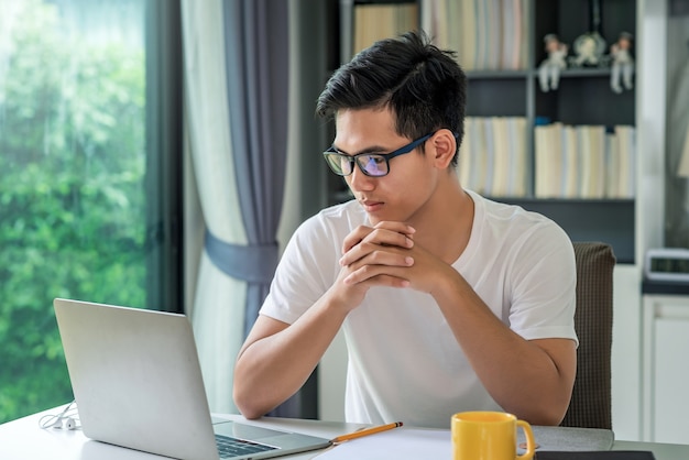 Vue de face jeune homme asiatique travaillant à domicile en regardant l'ordinateur portable.