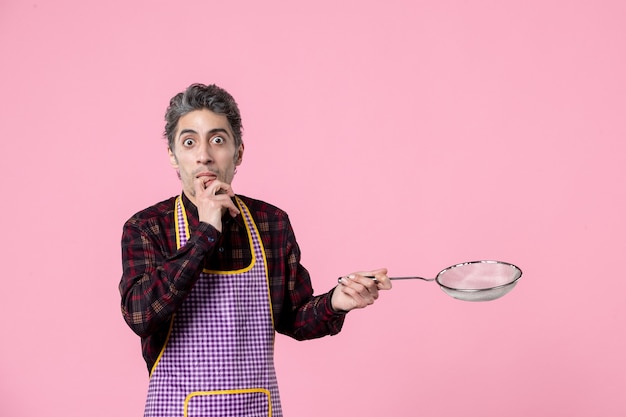 Photo vue de face jeune homme en cape holding tamis sur fond rose ouvrier cuisinier profession mari cuisine couleur alimentaire horizontale