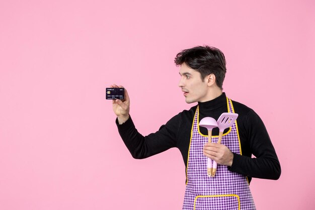 Vue de face jeune homme en cape tenant une carte bancaire et des cuillères sur fond rose travail travailleur couleur profession uniforme argent chef cuisine horizontale cuisine