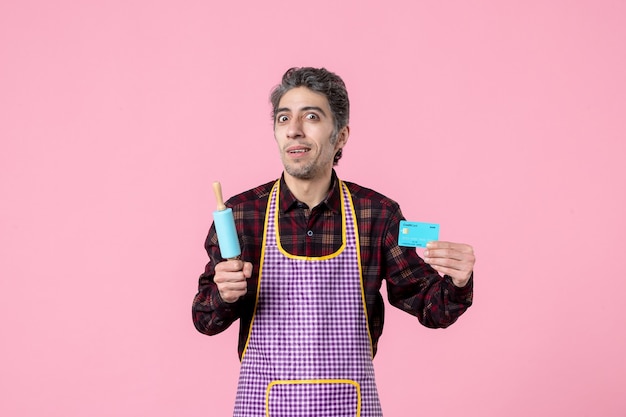 vue de face jeune homme en cape tenant un petit rouleau à pâtisserie et une carte bancaire sur fond rose argent profession mari cuisson travail travailleur cuisine horizontale