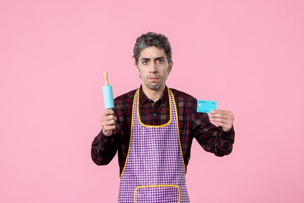 vue de face jeune homme en cape tenant un petit rouleau à pâtisserie et une carte bancaire sur fond rose profession mari travailleur cuisine horizontale uniforme travail de cuisine